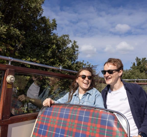 A couple in their early thirties in sunglasses laugh on a vintage, open-topped bus.