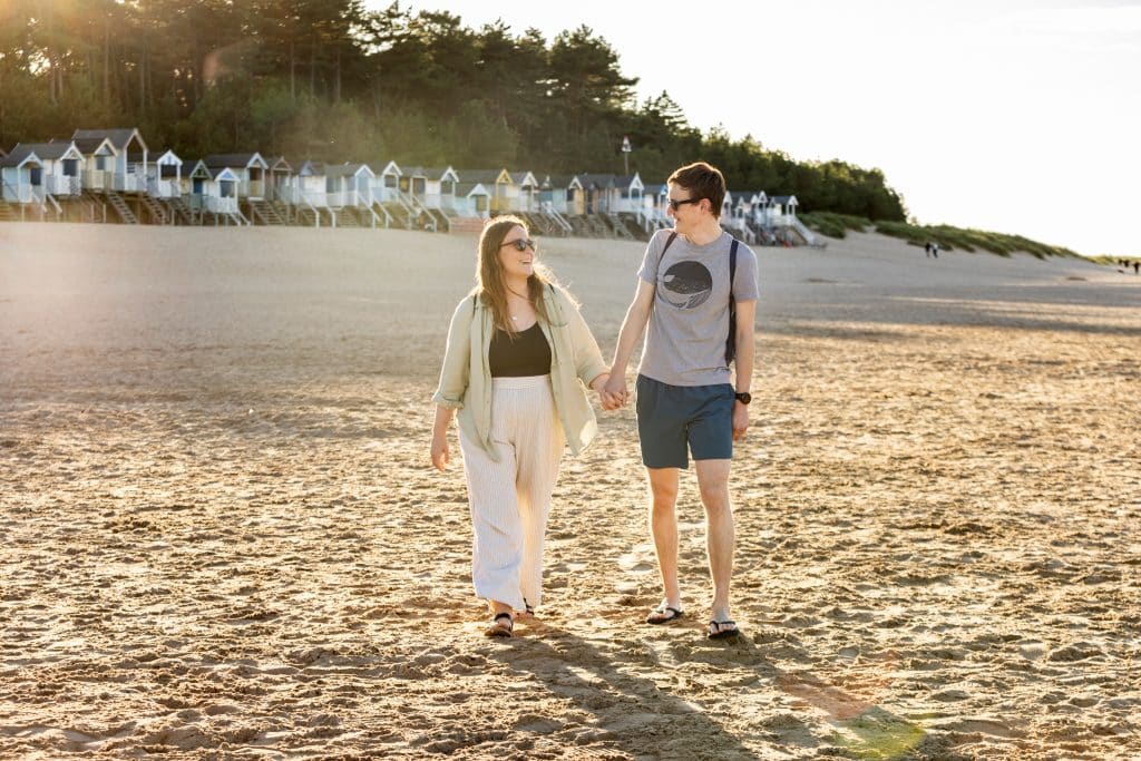 A couple walk along sandy Wells Beach in Norfolk, with the beach huts in the background.