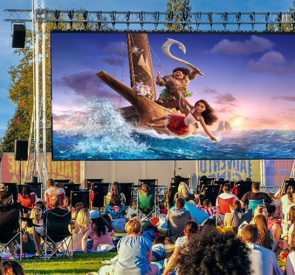 A large open air film screen showing a scene from Moana with families watching it on camping chairs.