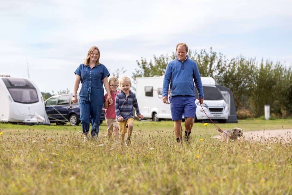Two parents and their young sons and dog walk through the grass on Pinewoods' touring park, with a caravan, car and motorhome in the background.