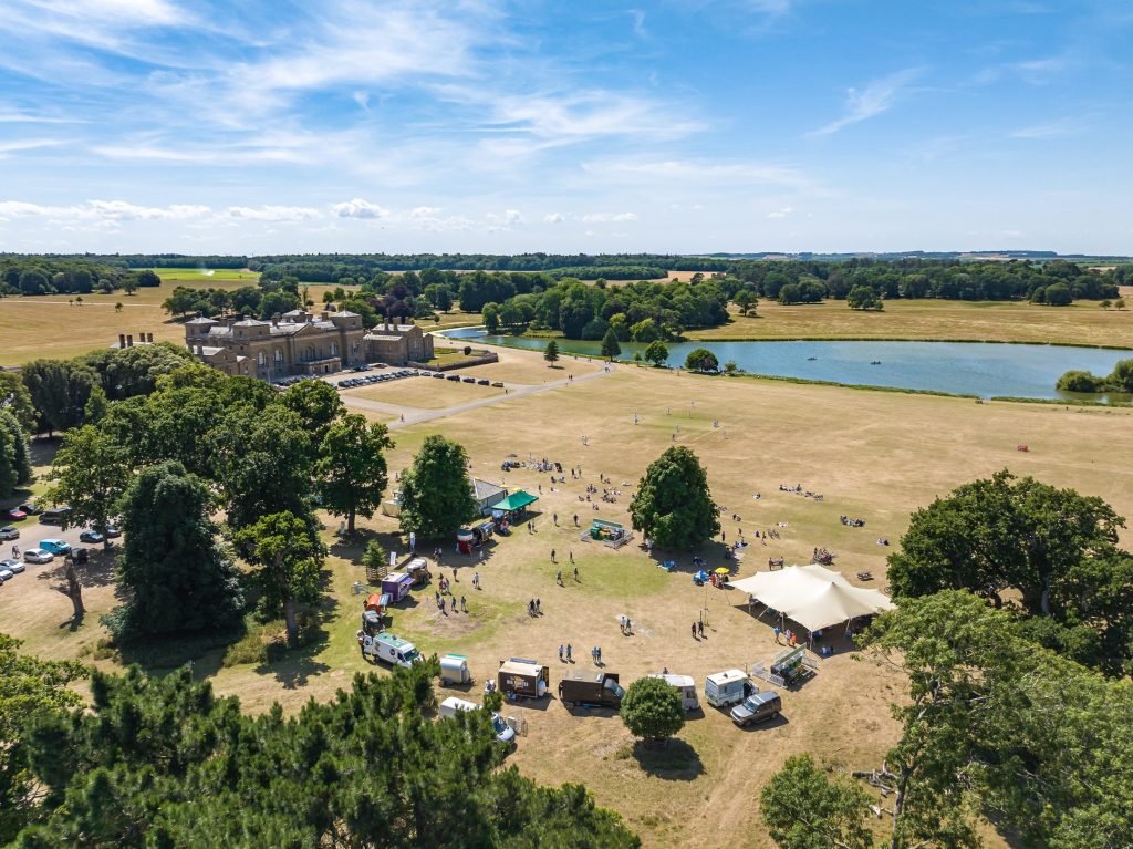 An aerial photo of Holkham Park and lake, with food trucks, a sail tent marquee and benches.