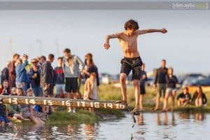 A white boy in dark shorts sliding along a wooden pole over the harbour at Blakeney.