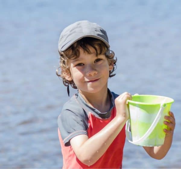 A young boy in a cap and tshirt holds a bucket by the sea.