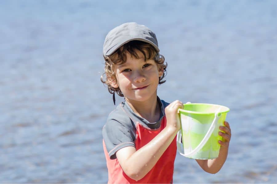 A young boy in a cap and tshirt holds a bucket by the sea.