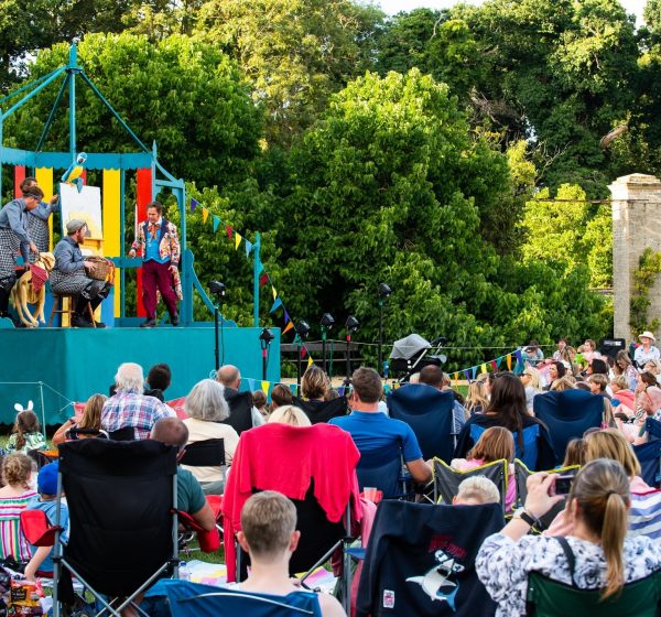 A crowd sit on camping chairs watching performers on an outdoor stage