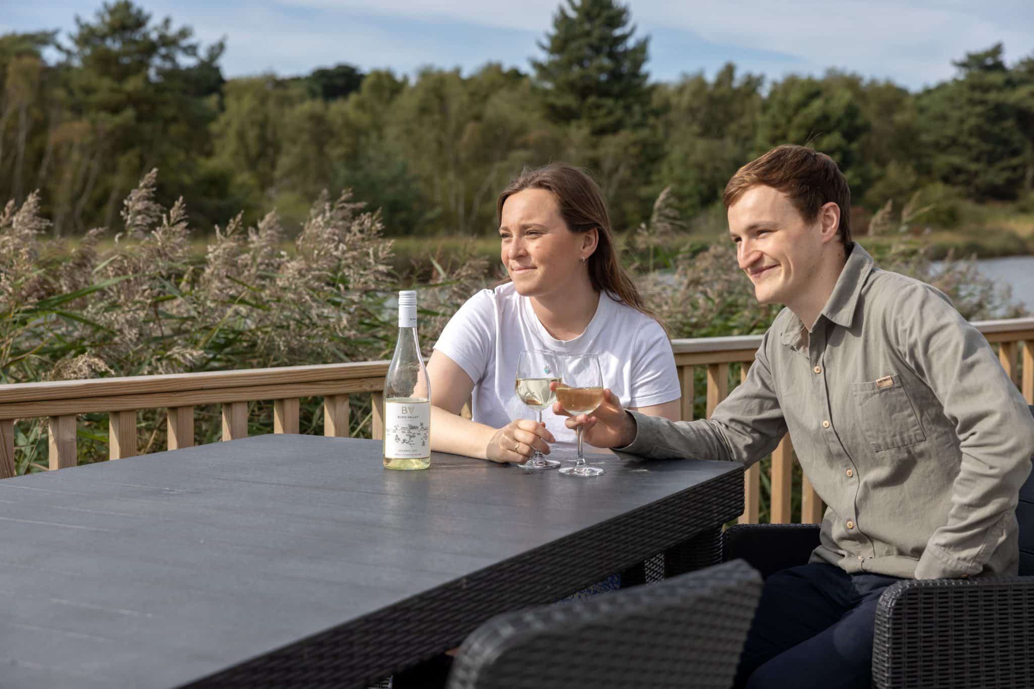 A couple sit at a table on the decking of a holiday lodge, enjoying a glass of white wine each and looking at the lake and trees