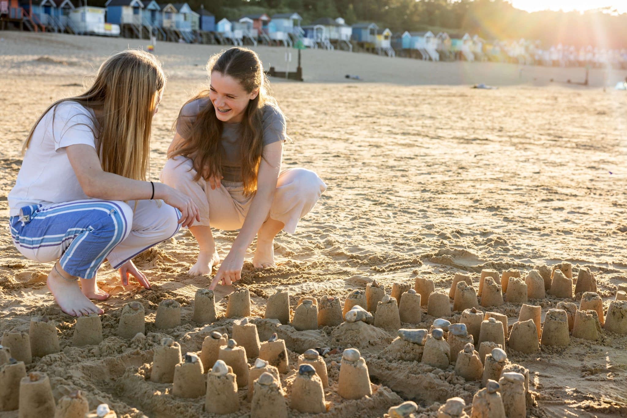 Two teenage girls laugh while crouching on the sand next to a large group of sandcastles on Wells Beach