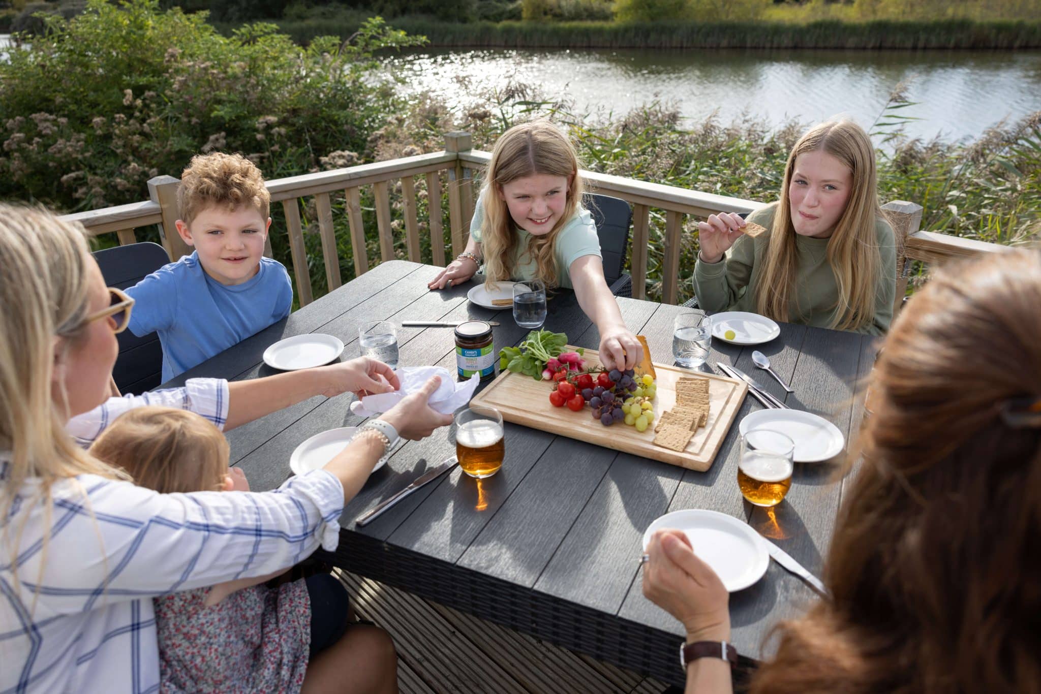 A family sit and eat nibbles on the decking of a holiday lodge at Pinewoods, overlooking the lake