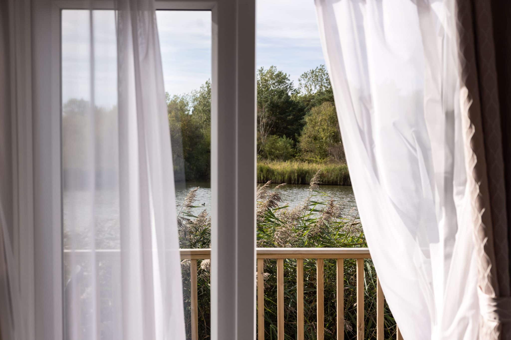 Curtains blow in the breeze with a view through a french door out to the decking and lake view.