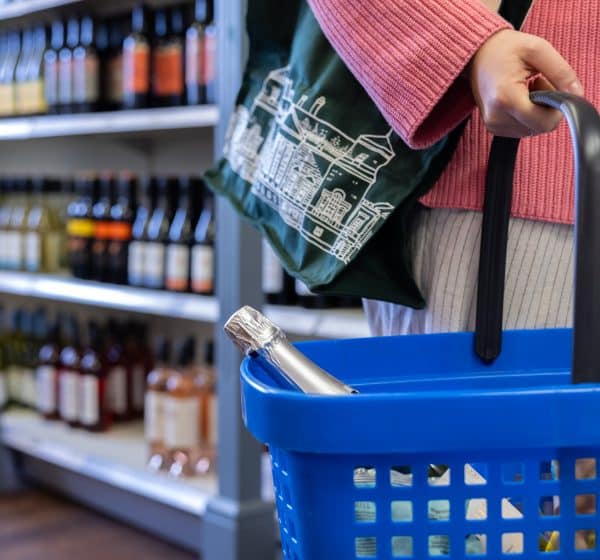 A person holds a shopping basket with a bottle of Prosecco sticking out