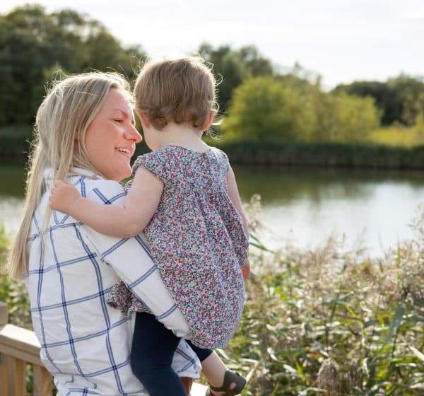 A mum holds her toddler as they look out at a view of a lake and trees at Pinewoods Holiday Park