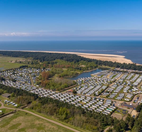 An aerial photo showing the Pinewoods Spoonbill Drift touring park with caravans, campervans and motorhomes pitched up. Behind is the caravan park full of static caravans surrounded by trees, then Wells Beach and the sea in the background