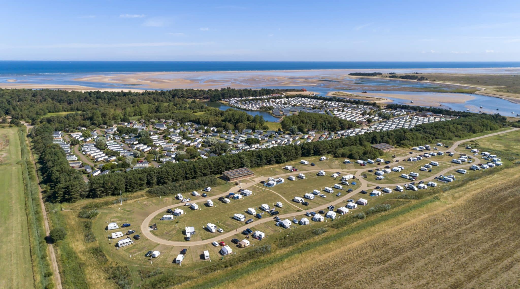 An aerial photo showing the Pinewoods Spoonbill Drift touring park with caravans, campervans and motorhomes pitched up. Behind is the caravan park full of static caravans surrounded by trees, then Wells Beach and the sea in the background
