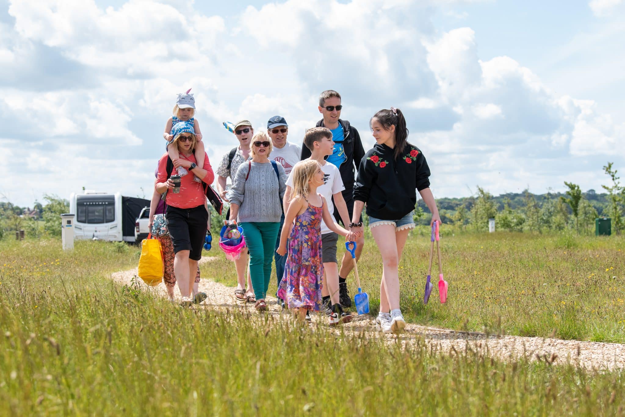 A large family in summer clothing walk along a path in a touring park, holding buckets, spades and beach equipment