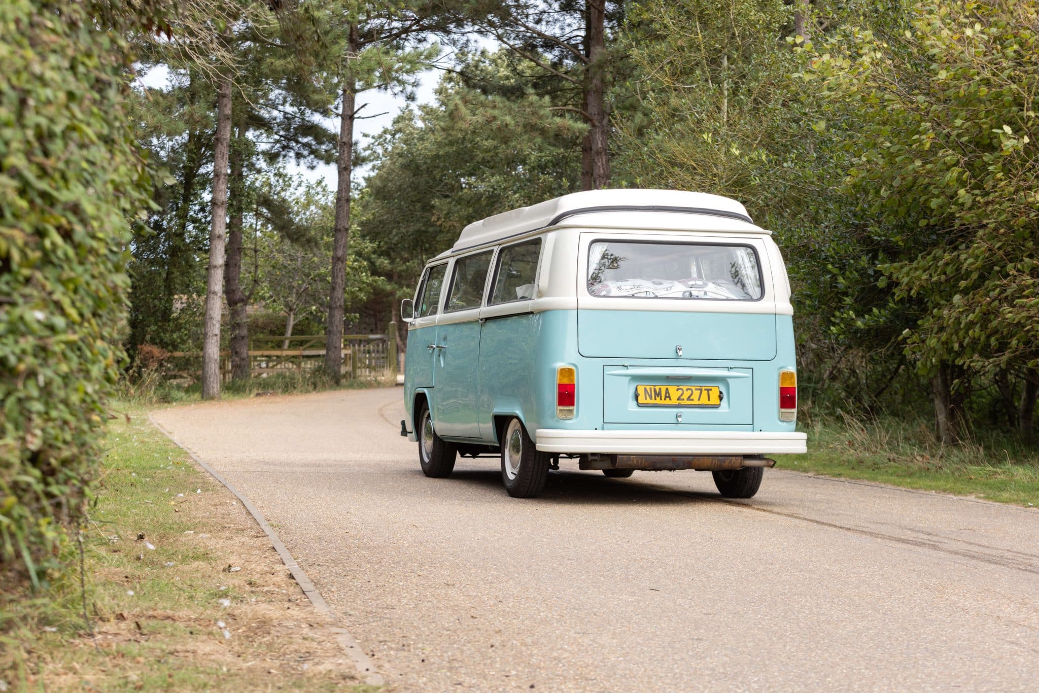 A vintage, pale blue and white Volkswagen campervan driving away from camera down a lane between trees