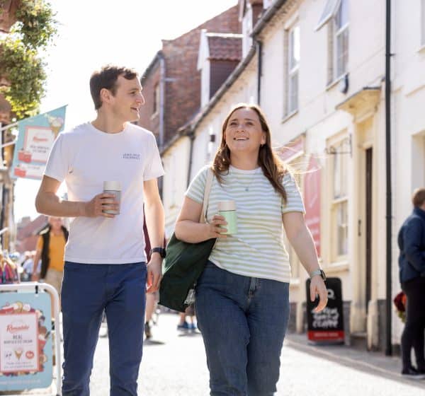 A couple walk with coffee cups down a sunny Staithe Street.