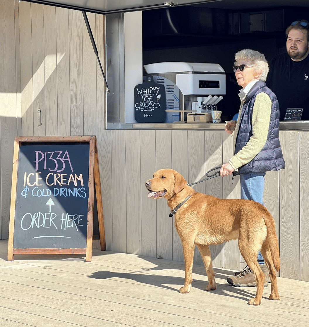 A lady stands with her labrador outside the hatch at Wells Beach Café. A sign advertises pizza and ice cream.