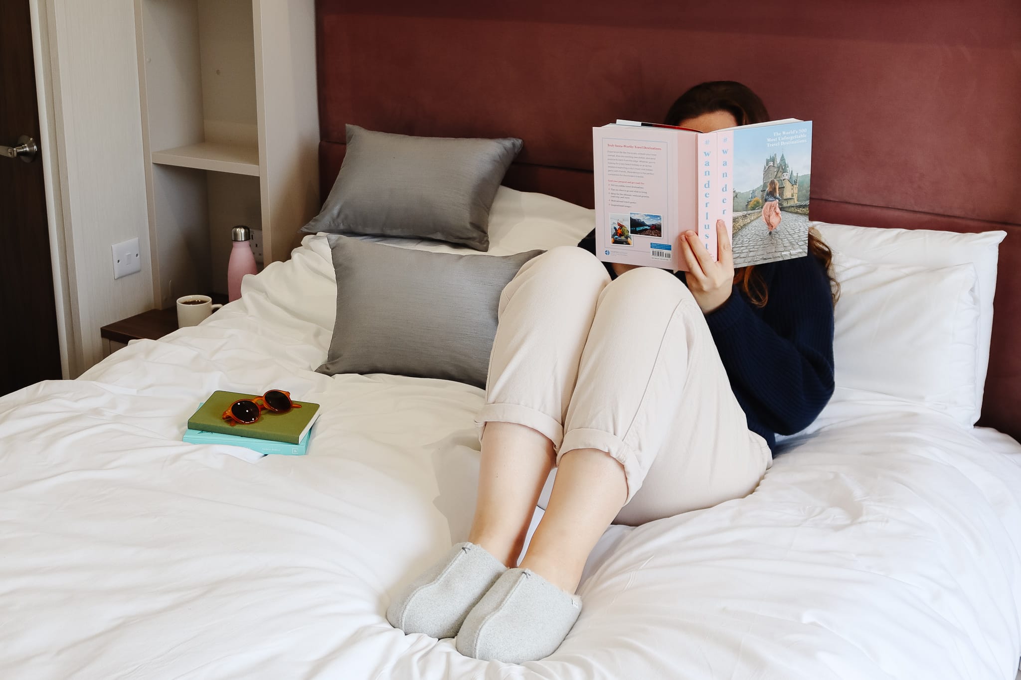 A woman reads a book on a bed, wearing a jumper, jeans and slippers.