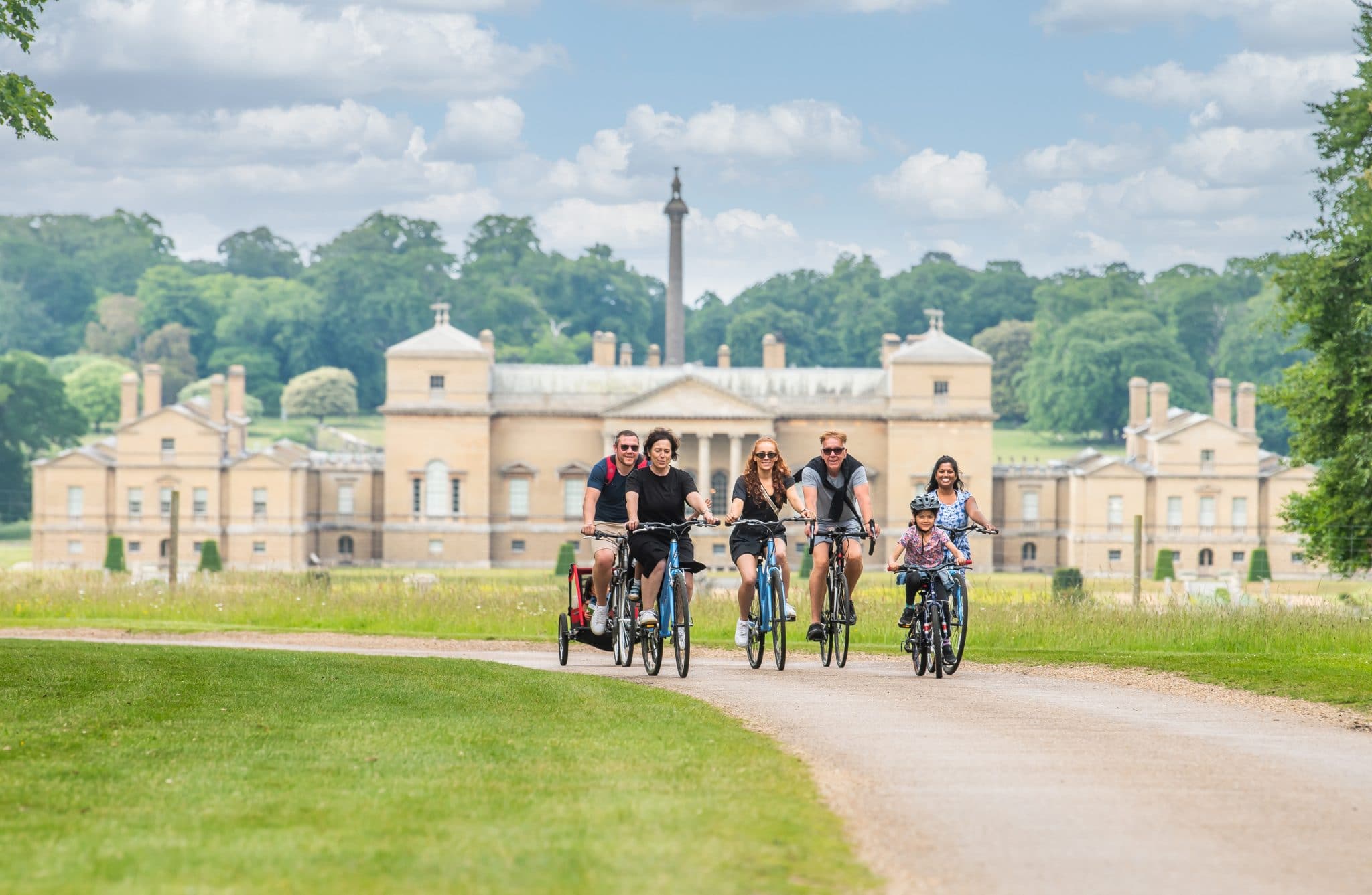 A group of adults and children cycle through Holkham Park, with Holkham Hall in the background.